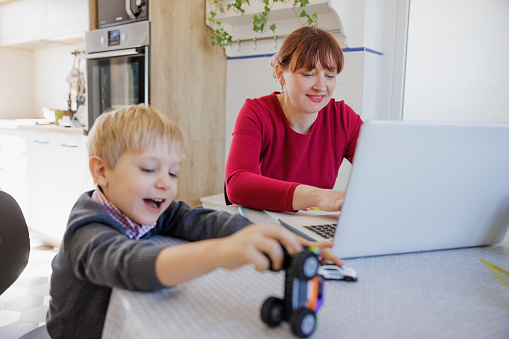 Smiling woman sitting at the table and working on laptop, her toddler making noise with toy truck and distracting her