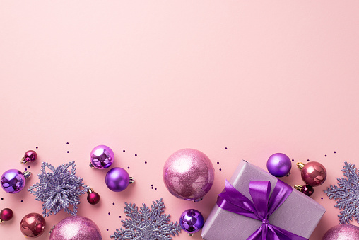 New Year concept. Top view photo of lilac present box with ribbon bow pink and violet baubles snowflake flower ornaments and sequins on isolated light pink background with copyspace