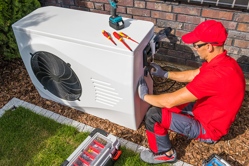Professional Middle Aged HVAC Technician in Red Uniform Repairing Modern Heat Pump Unit. House Heating and Cooling System Theme.
