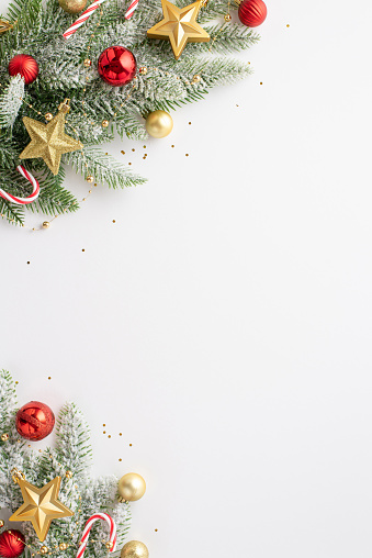 Christmas concept. Top view vertical photo of pine branches in snow decorated with red baubles gold star ornaments candy canes and confetti on isolated white background with empty space