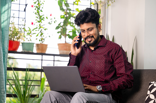 Young Asian Indian Businessman working on laptop and using smartphone at home with plants and greenery in background.