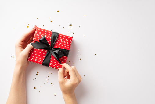 Black friday concept. First person top view photo of girl's hands untying ribbon bow on red giftbox over gold glowing confetti on isolated white background