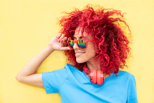 close-up profile of a smiling young Latin woman with red afro hair wearing a blue T-shirt and sunglasses on a yellow background, red headphones around her neck