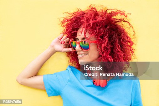 istock close-up profile of a smiling young Latin woman with red afro hair wearing a blue T-shirt and sunglasses on a yellow background, red headphones around her neck 1438147656