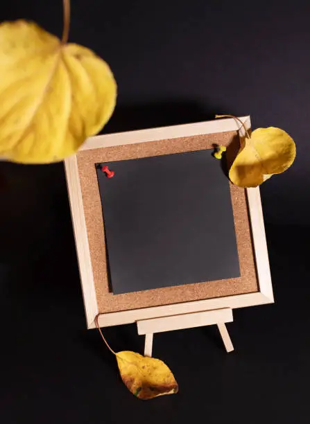 Photo of Yellow fall leaves and small decorative wooden easel with empty black sheet on black background.
