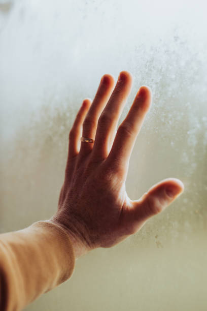 istock Old woman's hand on a window wearing a marriage ring - husband loss concept 1438148191