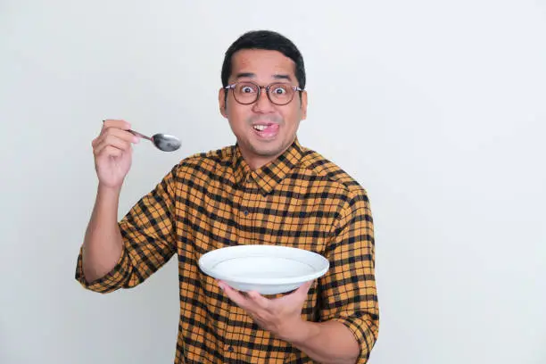 Adult Asian man holding empty dinner plate and spoon with funny expression