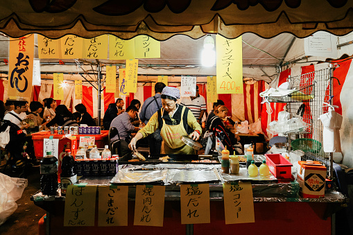 Tokyo, Japan - November 1, 2022 : Local Japanese street food at night market in Tokyo, Japan.  woman grilling yakitori pork and chicken at street food vendor market in the Japanese winter festival.