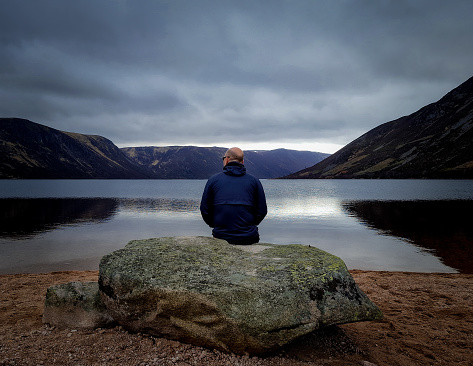 Man sits by himself in quiet contemplation on a dark day at loch Muick near Ballater