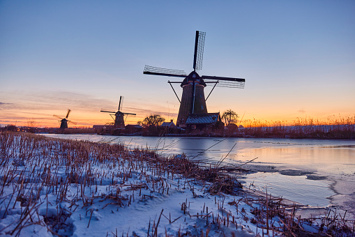 Several windmills in winter at the Kinderdijk, Holland. The water is frozen and all the land is covered with snow. There is a bridge over get water. The sun has just set.