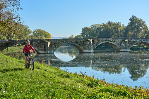 nice senior woman riding her electric mountain at river Arno near Arezzo, with famous Ponte Buriano in background, Arezzo,Tuscany , Italy