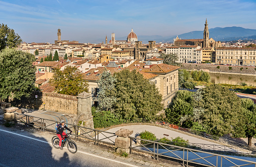 cheerful senior woman cycling with her electric mountain bike in downtown of Florence with famous bridge of ponte Vecchio in background, Tuscany, Italy