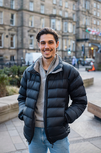 A young man spending the day in Newcastle Upon Tyne. He is standing on a paved area in the city centre with buildings behind him. He is looking at the camera and smiling.