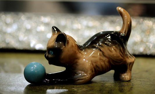 A closeup of a ceramic cat statue playing with a ball on the table with a blurry background