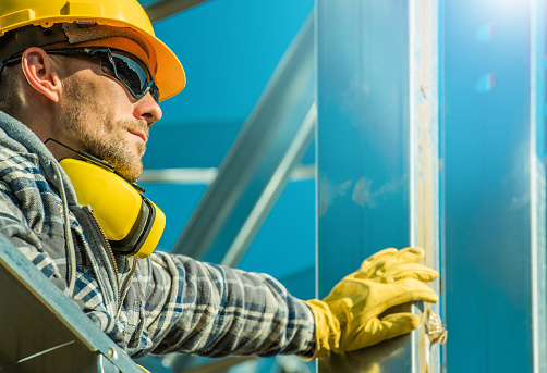 Caucasian Construction Industry Worker Wearing Yellow Hard Hat and Sunglasses Staying Next to Aluminium Building Frame.