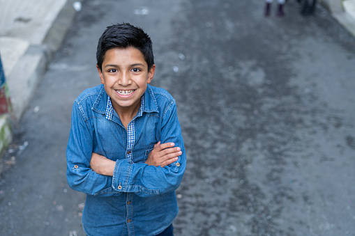 Latino boy of average age of 10 years casually dressed in a jeans shirt stands in the street of his neighborhood looking at the camera that portrays him while smiling