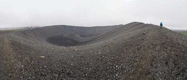 Two women hiking the edge of the Hverfjall volcanic crater on cloudy day.  Hverfjall, is one of the best preserved circular volcanic craters in the world and it is possible to walk around and inside it. Panorama.