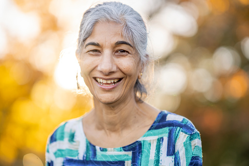 A senior woman of East Indian decent, poses for a portrait while outside on a cool fall day.  She is dressed casually in a printed blue shirt and has her hair pulled back as she smiles warmly for the camera.