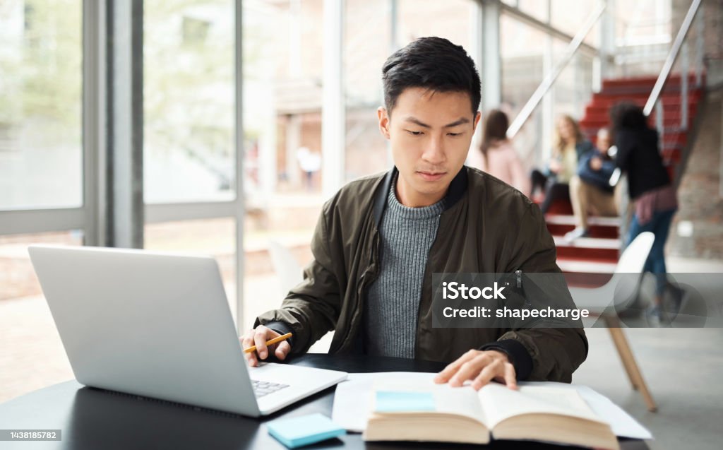 Laptop, reading book and asian student studying in university hall, campus library or workspace for education, exam and knowledge learning. Japanese man research on pc with language report writing Student Stock Photo