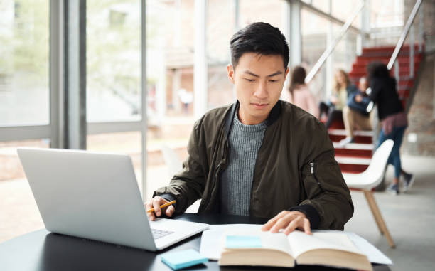 Laptop, reading book and asian student studying in university hall, campus library or workspace for education, exam and knowledge learning. Japanese man research on pc with language report writing stock photo