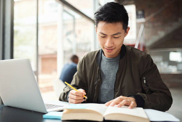 istock Asian student man, writing notebook with laptop in education learning at study hall desk with notes. Young studying guy with note, books and computer at table for exam success at university college 1438185801