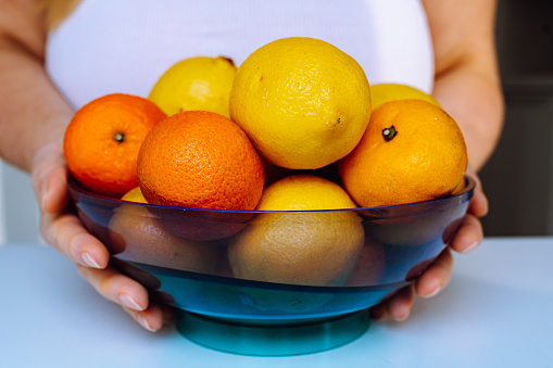 Close-up female hands holding blue glass bowl with lemons, tangerines and oranges on white table selective focus