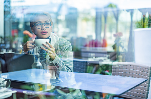 View through the window of a young woman sitting in cafe and holding cup of coffee