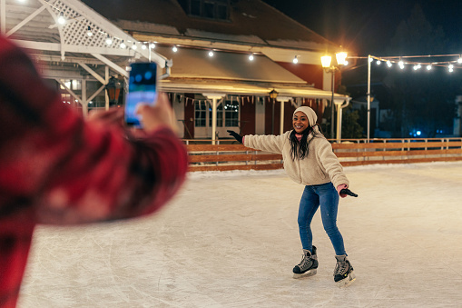 Romantic couple taking photo on the ice rink in the winter. The couple is wearing casual winter warm clothes. Woman posing while her boyfriend taking photo of her