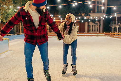 Young man ice skating outdoors, giving assistance to his african partner and teaching her how to ice skate.