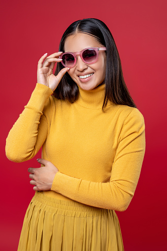 Smiling cute young fashionista in the yellow dress touching the rim of her sunglasses and looking ahead