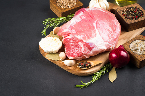 Boneless raw pork shoulder ready for roasting with seasonings and vegetables over dark concrete background with copy space for text. Organic meat for main dish recipe