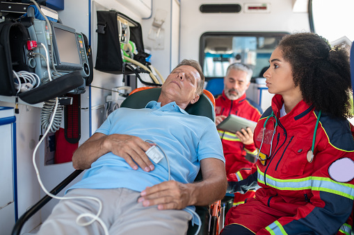 Serious focused ambulance doctor sitting beside an unconscious man lying on the gurney and looking at the ECG monitor