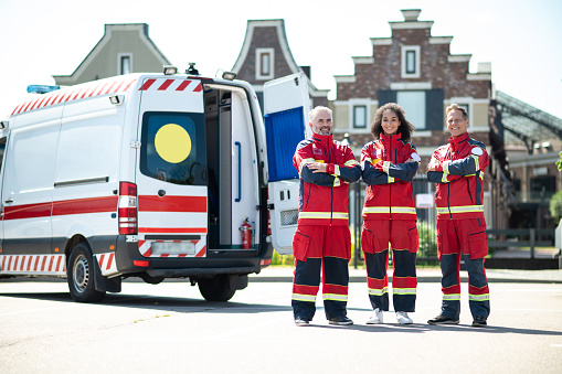 Cheerful team of modern paramedics dressed in the red uniforms standing beside the EMS vehicle