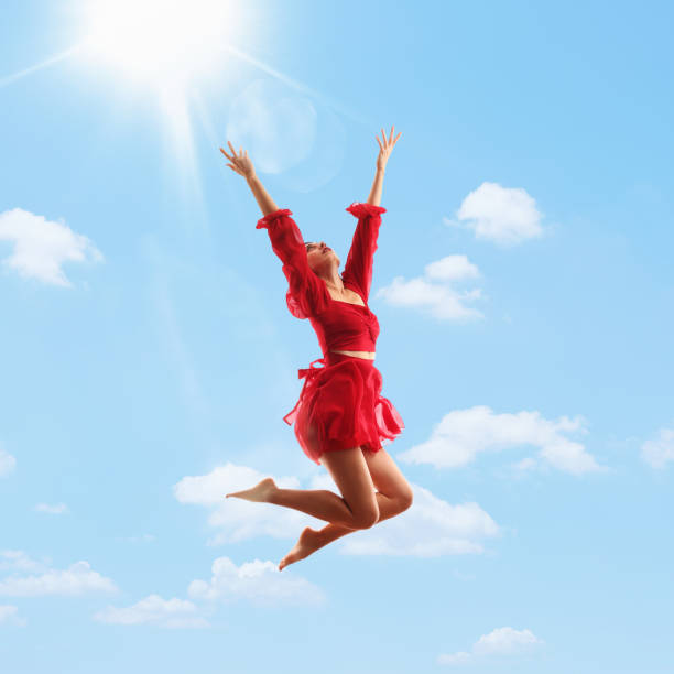 jovem de vestido vermelho pulando no ar, braços levantados, céu azul e sol como fundo - women jumping bouncing spring - fotografias e filmes do acervo