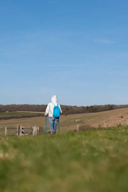 A woman wearing a white coat is walking with blue sticks and carrying a blue bag. She is seen from the back, her coat's hoodie is on and she's wearing jeans. The area is Seven Sisters, England.
