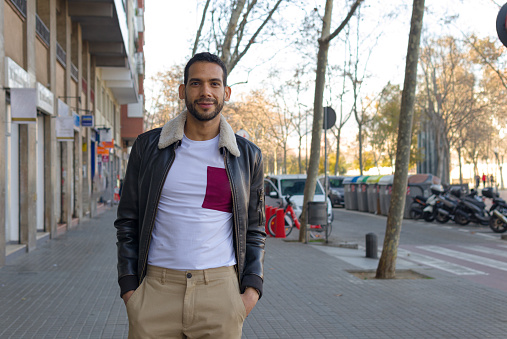 three quarter length shot of a latin man standing on a street and looking at camera with hands in pockets