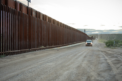 The international border wall between San Diego, California and Tijuana, Mexico, with an approaching U.S. Border Patrol vehicle on a nearby hill.