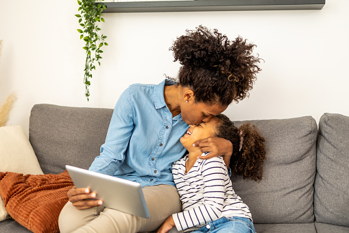 Mother and daughter sitting on the sofa. Mother is holding digital tablet and kissing her daughter in the forehead