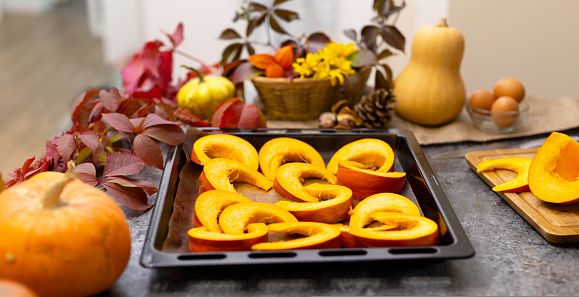 Pieces of orange pumpkin on a baking sheet. On the background lie different pumpkins and beautiful autumn leaves. The table is decorated for Thanksgiving.