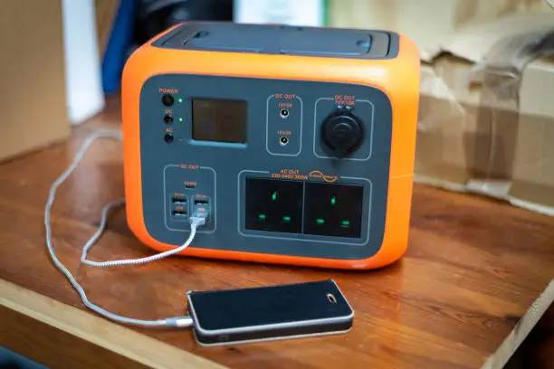 Portable power station solar electricity generator with mobile phone plugged in to charge. Wireless charging, lithium battery backup for power outage emergencies or outdoor travel camping.