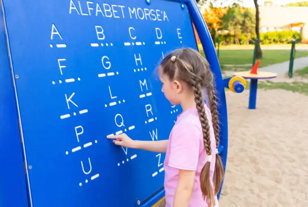 A little girl 7-8 years old with long hair dressed in a pink T-shirt and shorts is studying Morse code on a blackboard in the playground.