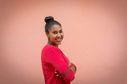 Side view of a young beautiful black woman smiling while looking at camera wearing red blouse in front of a coral color background