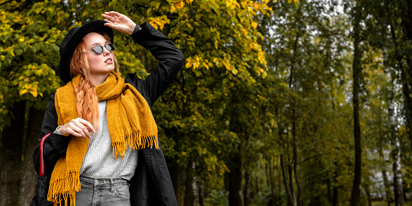 Portrait of a red haired woman in black glasses and a black hat in an orange scarf on the background of a park in early autumn.