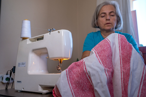 55-60 years old old woman with white hair. She is wearing a blue shirt inside the house. She sews with a machine at home. She is highly concentrated and careful. waist up