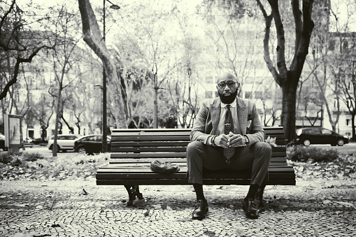 Black and white shot with an elegant bearded bald African man entrepreneur in a fashionable tailored formal suit with a necktie and eyeglasses sitting on a park bench on an autumn day, a road behind