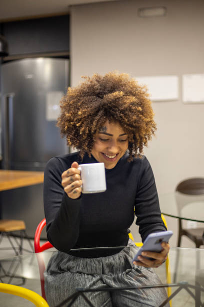 Black woman using cellphone and drinking coffee in office break area