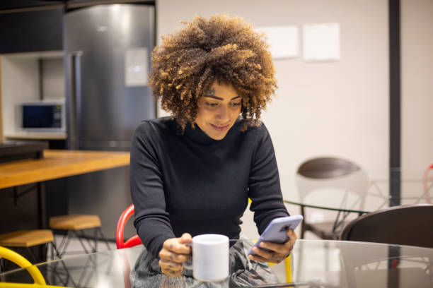 istock Black woman using cellphone and drinking coffee in office break area 1438254166