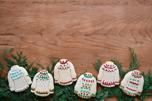 Homemade ugly sweater Christmas cookies framing bottom of wooden background with green cedar branches and copy space