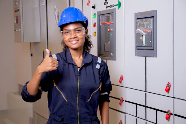 Electrical young asian woman engineer examining maintenance cabinet system electric and smile in control room at industrial factory, technician or electrician inspection power distribution. stock photo