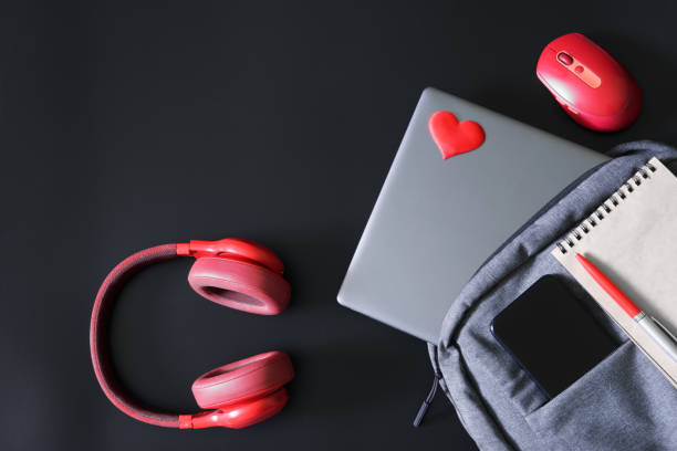 Gray trendy laptop backpack with a mobile phone, a notepad, a pen and a laptop on a black background next to red headphones and a mouse. Soft laptop sticker in the shape of a heart. Close-up